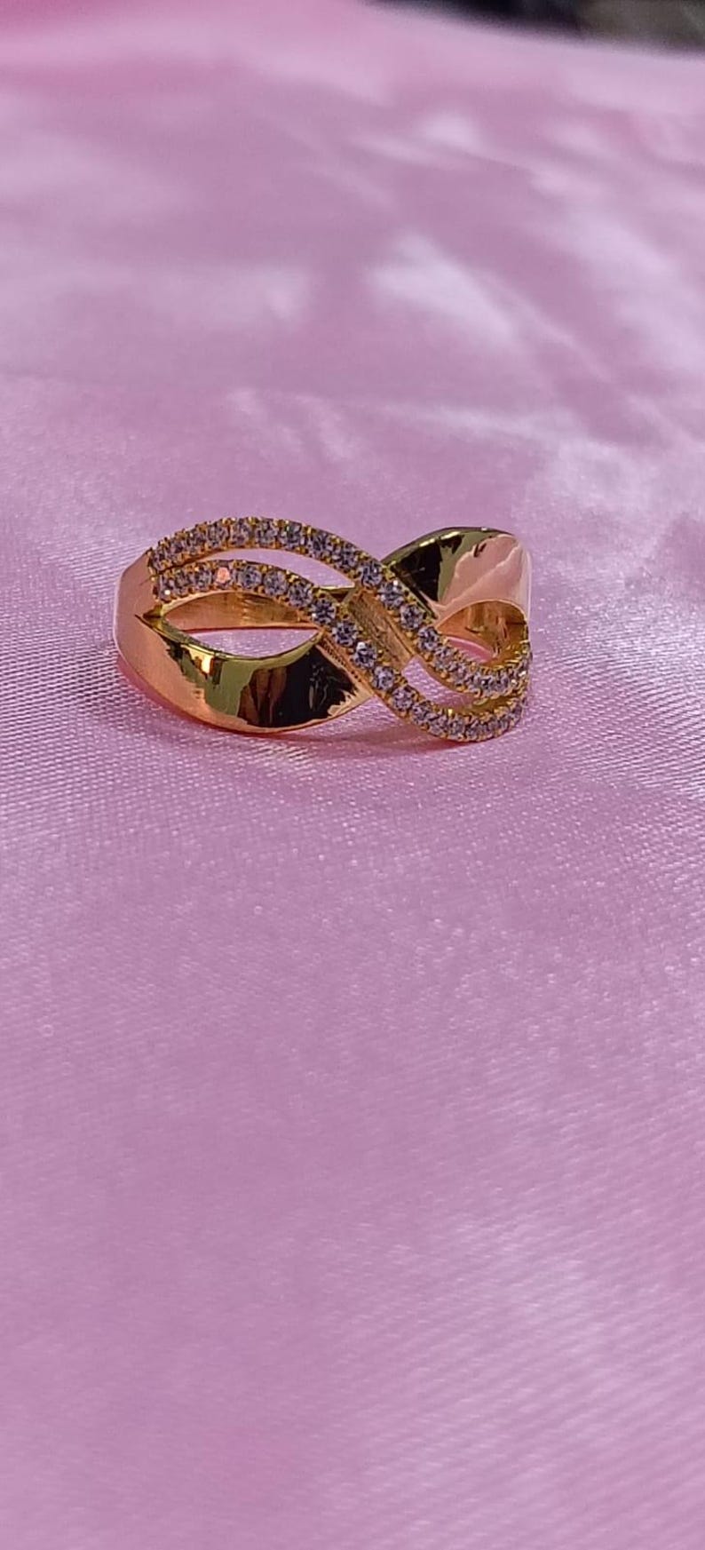 May include: A gold-colored ring with an infinity symbol design. The ring is adorned with small, clear gemstones. The ring is set against a pink background, creating a contrast that highlights the ring's details.