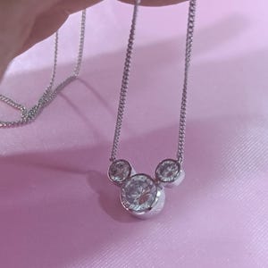 Op de afbeelding: Een zilverkleurige ketting met drie heldere, ronde edelstenen gerangschikt in de vorm van een Mickey Mouse-hoofd. De ketting heeft een fijne ketting en wordt gepresenteerd tegen een zachtroze achtergrond. De edelstenen schitteren.