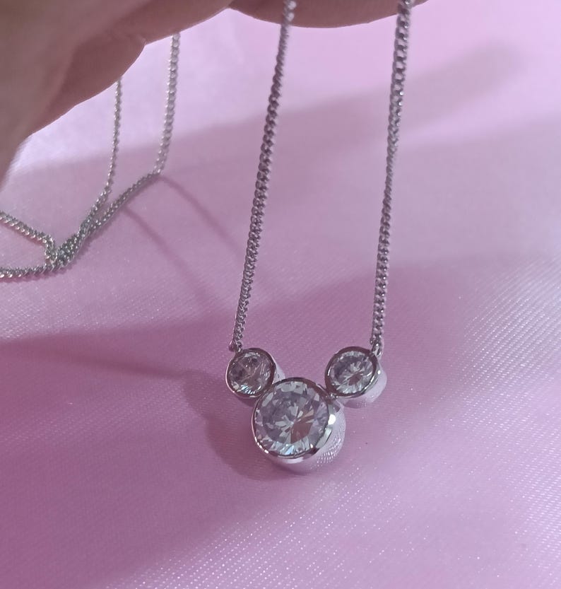 Op de afbeelding: Een zilverkleurige ketting met drie heldere, ronde edelstenen gerangschikt in de vorm van een Mickey Mouse-hoofd. De ketting heeft een fijne ketting en wordt gepresenteerd tegen een zachtroze achtergrond. De edelstenen schitteren.