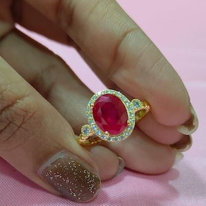 May include: A gold ring featuring a large oval-cut ruby center stone surrounded by a halo of small, clear gemstones. The ring has additional small gemstones on each side of the main stone, set on a gold band.