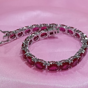 May include: A silver-toned tennis bracelet featuring a continuous row of oval-cut, vibrant pinkish-red gemstones set in individual metal prongs. The bracelet includes a secure box clasp mechanism and is arranged in a circular shape on a smooth surface.
