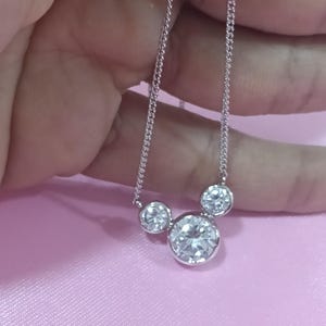 Op de afbeelding: Een zilveren ketting met drie ronde, heldere edelstenen. De grootste edelsteen staat centraal, met twee kleinere stenen aan weerszijden. De ketting wordt getoond tegen een roze achtergrond.