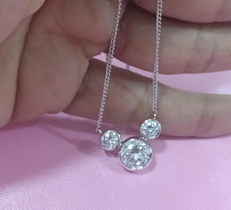 Op de afbeelding: Een zilveren ketting met drie ronde, heldere edelstenen. De grootste edelsteen staat centraal, met twee kleinere stenen aan weerszijden. De ketting wordt getoond tegen een roze achtergrond.