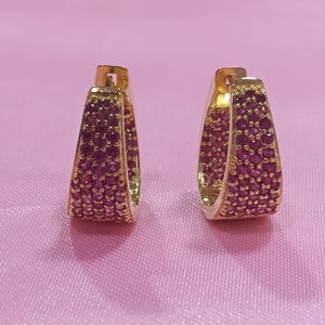 May include: A pair of gold hoop earrings featuring a wide, tapered design. The front surface of each hoop is densely encrusted with numerous small, round, deep pink gemstones arranged in a structured, multi-row pattern across the metal frame.