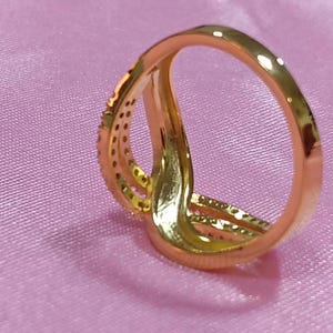 May include: A gold-colored ring with a unique design, featuring multiple bands that intertwine. One band is adorned with small, sparkling stones. The ring is set against a soft, pink background.