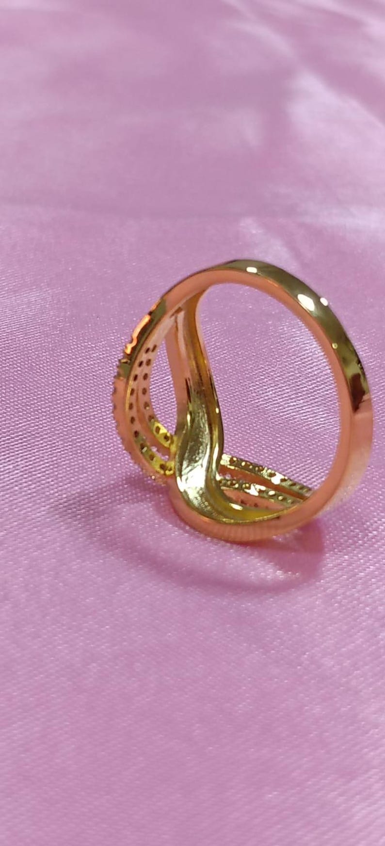 May include: A gold-colored ring with a unique design, featuring multiple bands that intertwine. One band is adorned with small, sparkling stones. The ring is set against a soft, pink background.