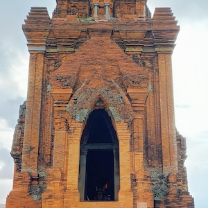 May include: A tall, ancient brick tower with an arched entrance and intricate carvings. The structure is a reddish-brown color and stands against a cloudy sky. The tower is surrounded by green grass.