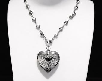 May include: A silver-toned necklace with a heart-shaped locket pendant. The chain features small heart-shaped links, and the locket is intricately detailed with a floral design. The necklace is displayed on a white mannequin.