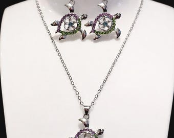May include: A silver-toned jewelry set featuring a turtle design. The set includes a necklace and a pair of earrings. The turtle pendants and earrings are adorned with purple, green, and blue gemstones, creating a sparkling effect. The jewelry is displayed on a white mannequin.