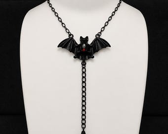 May include: A black lariat-style necklace featuring a central bat pendant with a small red accent on its chest. The bat has outstretched wings and is connected to a vertical chain drop that ends with a faceted black teardrop-shaped bead pendant.