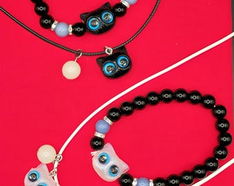 May include: Jewelry set featuring black beaded bracelets and necklaces with cat-shaped pendants. The cat pendants have blue eyes and are accented with light blue beads and silver-colored spacers. The background is red.