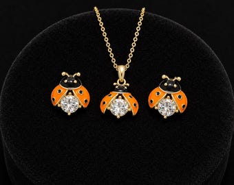 May include: A gold-toned jewelry set consisting of a necklace and two stud earrings. Each piece features a ladybug design with orange wings, black heads, and a central clear round faceted stone. The necklace includes a delicate gold chain.