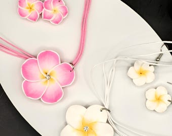 May include: A collection of floral jewelry, including earrings and necklaces. The earrings and one necklace feature pink and white plumeria flowers with yellow centers. The other necklace has a white flower with a yellow center. Each flower has a small, sparkling gem.