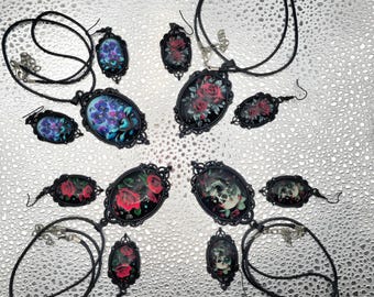 May include: An assortment of black pendant necklaces and earrings. The pendants feature floral and skull designs in oval frames. The jewelry is displayed on a textured silver surface. The floral designs include red roses and blue flowers.