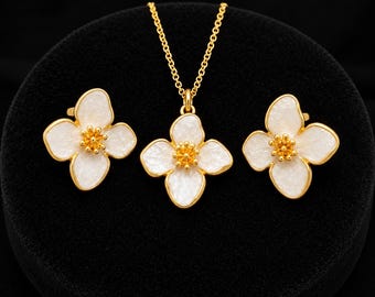 May include: A gold jewelry set consisting of a necklace and two matching earrings. Each piece features a four-petaled flower design with white enamel petals and a textured gold center. The necklace includes a delicate gold chain link.