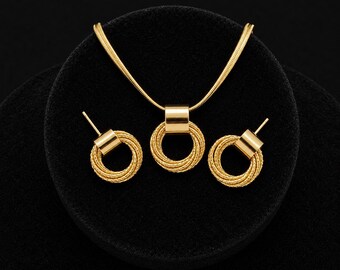 May include: A gold-toned jewelry set consisting of a necklace and two stud earrings. The necklace features a snake chain and a pendant with a smooth cylindrical bail holding a textured, multi-layered circular ring. The earrings match the pendant.