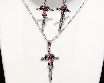 May include: A silver-toned jewelry set featuring a cross pendant necklace and matching earrings. Each piece is adorned with red rose accents. The jewelry is displayed on a white mannequin bust, showcasing the intricate details of the design.