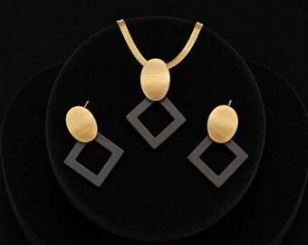 May include: A jewelry set consisting of a necklace and two earrings. Each piece features a brushed gold-tone oval component attached to a matte dark gray square frame. The necklace includes a gold-tone herringbone chain connected to the pendant.