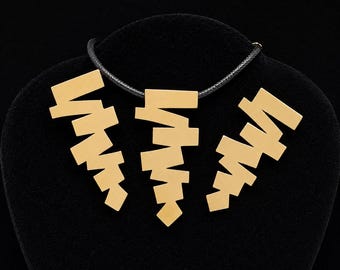 May include: A gold-toned necklace set featuring three geometric pendants with a stacked, zigzag-like arrangement of rectangular bars. The central pendant is suspended from a black cord, while the two matching side pendants mirror the design.