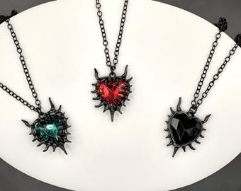 May include: Three heart-shaped pendant necklaces with black chains. Each pendant has a spiky, dark metal frame. The heart-shaped gems are emerald green, ruby red, and black. The necklaces are displayed on a white surface.
