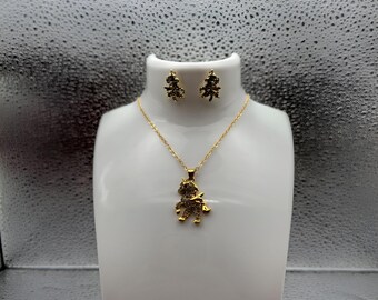 May include: A gold-colored jewelry set featuring a necklace and matching earrings. The necklace has a pendant with a figure design, and the earrings have a similar design. The set is displayed on a white bust against a silver background.