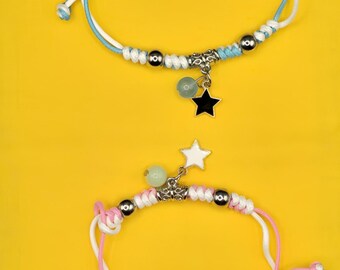 May include: Two handmade bracelets with adjustable cords. One bracelet is blue and white, with a black star charm. The other is pink and white, with a white star charm. Both have silver beads and a yellow background.