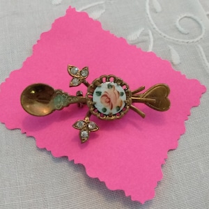 May include: A gold-toned metal brooch featuring a miniature spoon and heart-shaped handle design. The center displays a circular porcelain inlay with a painted pink rose motif, accented by two small leaf-shaped branches set with clear rhinestones.
