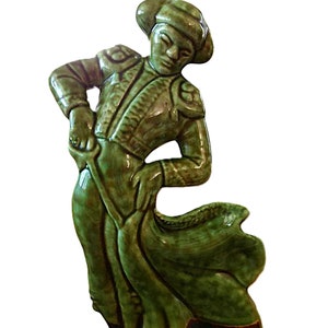 May include: A ceramic figurine of a matador in a green suit and hat, posed with a cape. The figurine has a glossy finish and is mounted on a dark brown base. The matador's stance suggests action and drama.