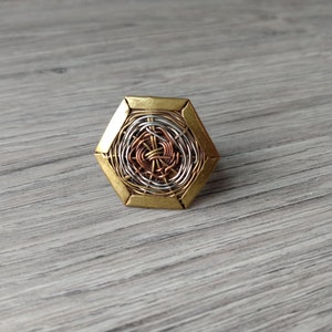 以下が含まれることがあります： 六角形の真鍮フレームを持つ、手作りの幾何学的なリング。リングの中央には、銀、銅、真鍮の複雑なワイヤーワークが施され、層状のテクスチャードデザインを作り出しています。ユニークなジュエリーです。