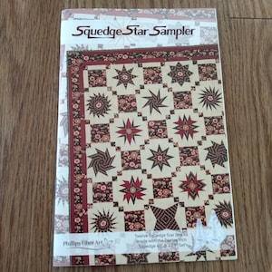 May include: A rectangular instructional booklet titled Squedge Star Sampler by Phillips Fiber Art. The cover features a quilt design with cream, dark red, and floral patterned blocks arranged in a grid, showcasing twelve star-shaped quilt blocks.