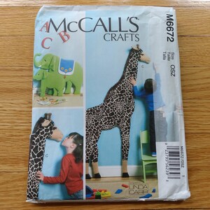 May include: McCall's Crafts sewing pattern M6672 for stuffed animal toys. The package features illustrations of a large giraffe with a black and white spotted pattern and a green elephant with a blue saddle. The text reads McCall's Crafts M6672.