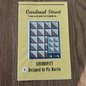 May include: A yellow booklet titled "Cardinal Street" with the quote "it seems we all needle little friendship sew...". The booklet features a quilt pattern design with geometric shapes in shades of brown, white, and navy. The word "SERENDIPITY" is also visible.