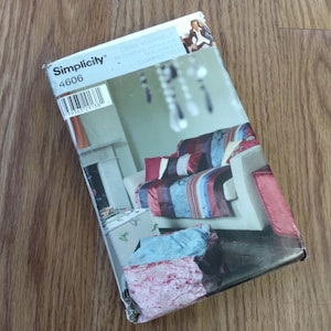 May include: A Simplicity sewing pattern envelope, model 4606, featuring a sofa with a striped cover in shades of red, blue, and burgundy. The envelope is on a wooden surface and includes the text "Deny Yourself Nothing... Completely".