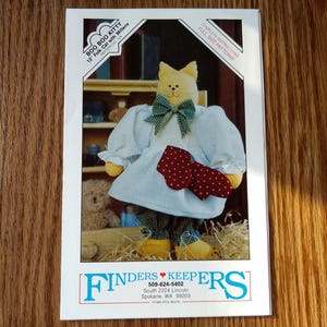 May include: A sewing pattern envelope titled Boo Boo Kitty 12 inch Folk Cat with Mittens by Finders Keepers. The cover features a yellow fabric cat wearing a white blouse, green plaid bow, and dark trousers, holding red mittens with white polka dots.