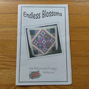 Peut inclure: Un livret en papier blanc intitulé Endless Blossoms avec une photographie centrale d'une courtepointe en patchwork multicolore aux motifs floraux dans des tons de violet, vert et bleu. Le bas inclut le texte The Patchwork Puppy Patterns.
