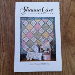 May include: A book cover with a quilt pattern in pastel shades and floral motifs. The title is "Shauna Case Quilters Clutter." The cover also shows a decorative birdhouse, cushions, and a box, with the word "SPRING" on a pillow.