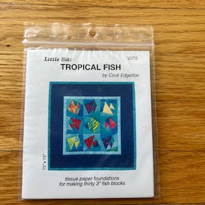 May include: A clear plastic package containing a Little Bits Tropical Fish quilt pattern by Cindi Edgerton. The white paper insert shows a blue-bordered quilt with colorful fish blocks, labeled 15" x 15" and tissue paper foundations for making thirty 3" fish blocks.