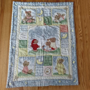 Peut inclure: Une couverture pour bébé matelassée avec des illustrations d'ours en peluche dans un motif patchwork. La bordure est bleu clair avec des étoiles et des lunes, avec des inscriptions comme "Les vœux se réalisent" et "Faites un vœu ce soir".