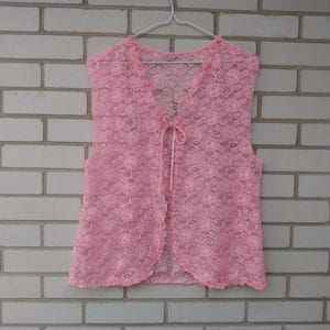 May include: A sleeveless pink lace waistcoat featuring an all-over floral pattern, a V-neckline, and a thin ribbon tie closure at the centre front. The garment is constructed from a sheer, lightweight lace fabric with scalloped edges along the hem.