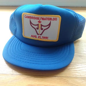 Könnte beinhalten: Blaue Trucker-Cap mit Mesh-Rückseite und gebogenem Schirm. Auf der Vorderseite befindet sich ein rechteckiger Aufnäher mit dem Text "CAMBRIDGE/WATERLOO", einem Bullen-Motiv und "AUG. 21, 1991".