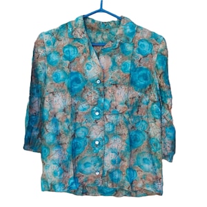 May include: A patterned blouse in teal, brown, and white, with a floral design. The blouse has a collared neckline and three-quarter sleeves. It is suspended from a blue clothes hanger.