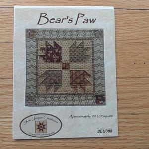 May include: A rectangular paper pattern titled Bear's Paw by Sew Unique Creations. The cover features a square quilt block with brown and tan patterned fabric in a paw print design. Text includes Approximately 20 1/2" square and SEU388.