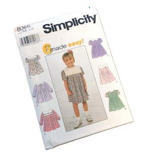 May include: A Simplicity sewing pattern envelope, number 8065, featuring six variations of a dress for children. The white envelope displays illustrations of dresses in floral, lavender, pink, and green fabrics, along with a barcode and the text 6 made easy.