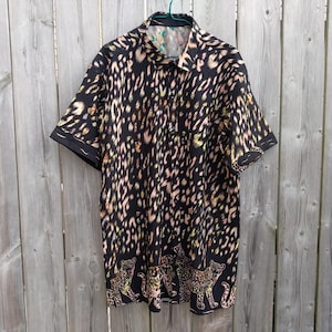 May include: A short-sleeved button-down shirt featuring a black base with an all-over leopard print pattern in shades of tan and white. The bottom hem displays a repeating graphic of standing leopards in a matching colour palette.