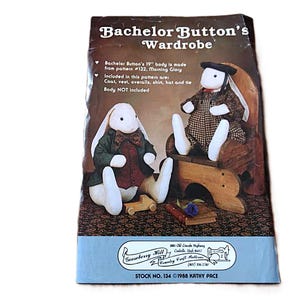 May include: A sewing pattern envelope titled Bachelor Button's Wardrobe, featuring two white stuffed rabbit dolls dressed in miniature clothing. One rabbit wears a green coat and red vest, while the other wears a brown plaid outfit and a black hat.