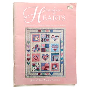 May include: A pink book cover with the title "A Celebration of Hearts" in elegant script. The cover features a quilt design with various heart patterns in squares, surrounded by a border of hearts. The authors' names are at the bottom.