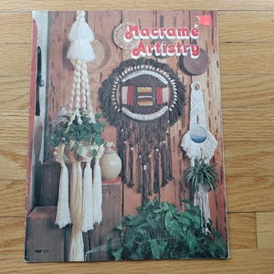 Peut inclure: Couverture d'un livre vintage intitulé "Macrame Artistry", présentant des suspensions de plantes en macramé, des tentures murales et un attrape-rêves décoratif. La couverture affiche un fond en bois rustique avec divers motifs en macramé et de la verdure.