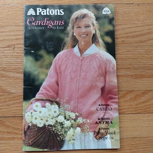 May include: A magazine cover showing a woman in a pink cardigan holding a basket of white flowers. The cover displays the title "Patons Cardigans" and text related to knitting.