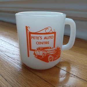 May include: A white milk glass mug featuring an orange graphic of a vintage car and a hanging sign with the text Pete's Auto Centre. The mug has a rounded body and a C-shaped handle on the right side, with the orange design printed on the front surface.