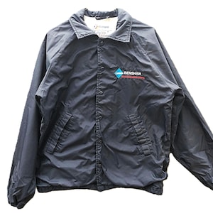 May include: A navy blue button-up jacket with a collar and two front pockets. The jacket features the text "BENSHAW" in red and a blue and white diamond logo on the left chest. The jacket is made of a lightweight material.